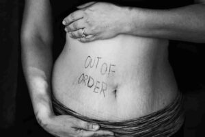 Black and white photo of a woman's torso with the words "Out of Order" written on the stomach. Her hands gently cradle the stomach.
